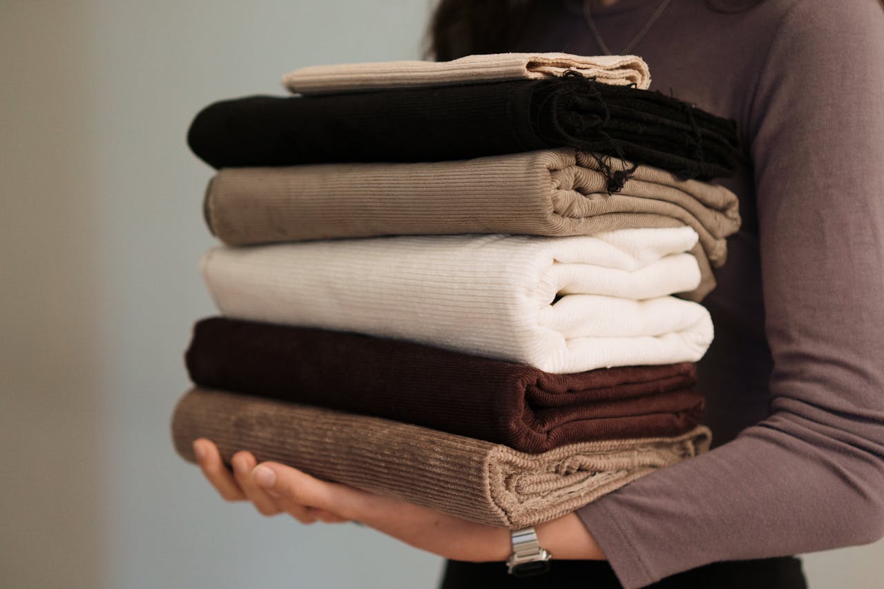 heros-img Close-up of hands holding a stack of folded neutral-toned fabrics, ideal for textile projects.