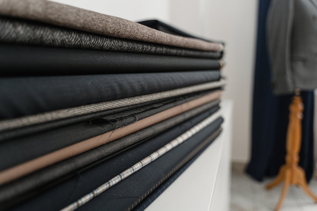 about-us A stack of various folded fabrics in a tailor shop, showcasing texture and color variety.
